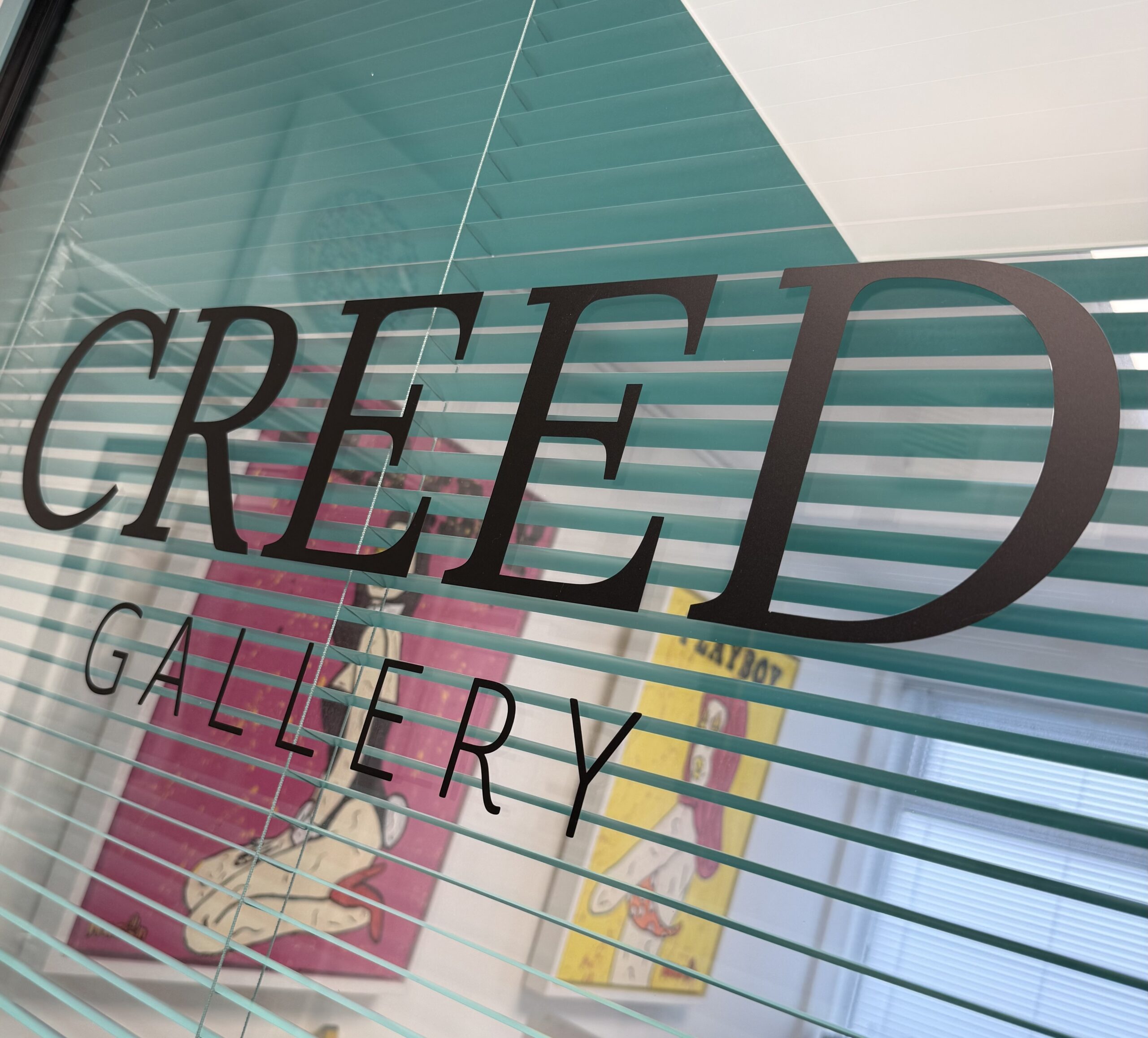 The Creed Gallery logo printed on glass with an aqua venetian blind in the background